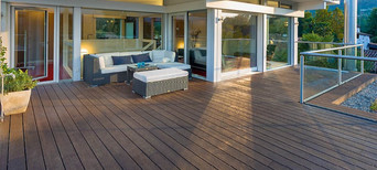 Moderne Terrasse mit dunklem Escheholz-Belag, stilvoller Rattan-Lounge mit weißen Polstern und direktem Zugang zum Wohnbereich.