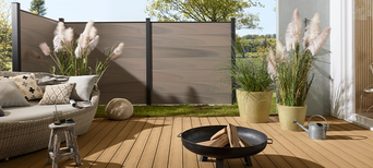 Runde Sonnenliege auf heller WPC-Terrasse mit Ziergräsern und stilvoller Feuerstelle. Im Hintergrund WPC-Zaun
