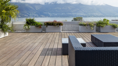 Große Terrasse mit Bambus-Holzdielen, modernen Rattanmöbeln, Pflanzen in Betonkübeln und Blick auf einen See mit Bergen.