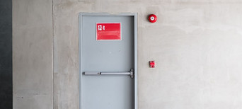 Graue Brandschutztür mit Fluchtwegsymbol und roter Alarmglocke.